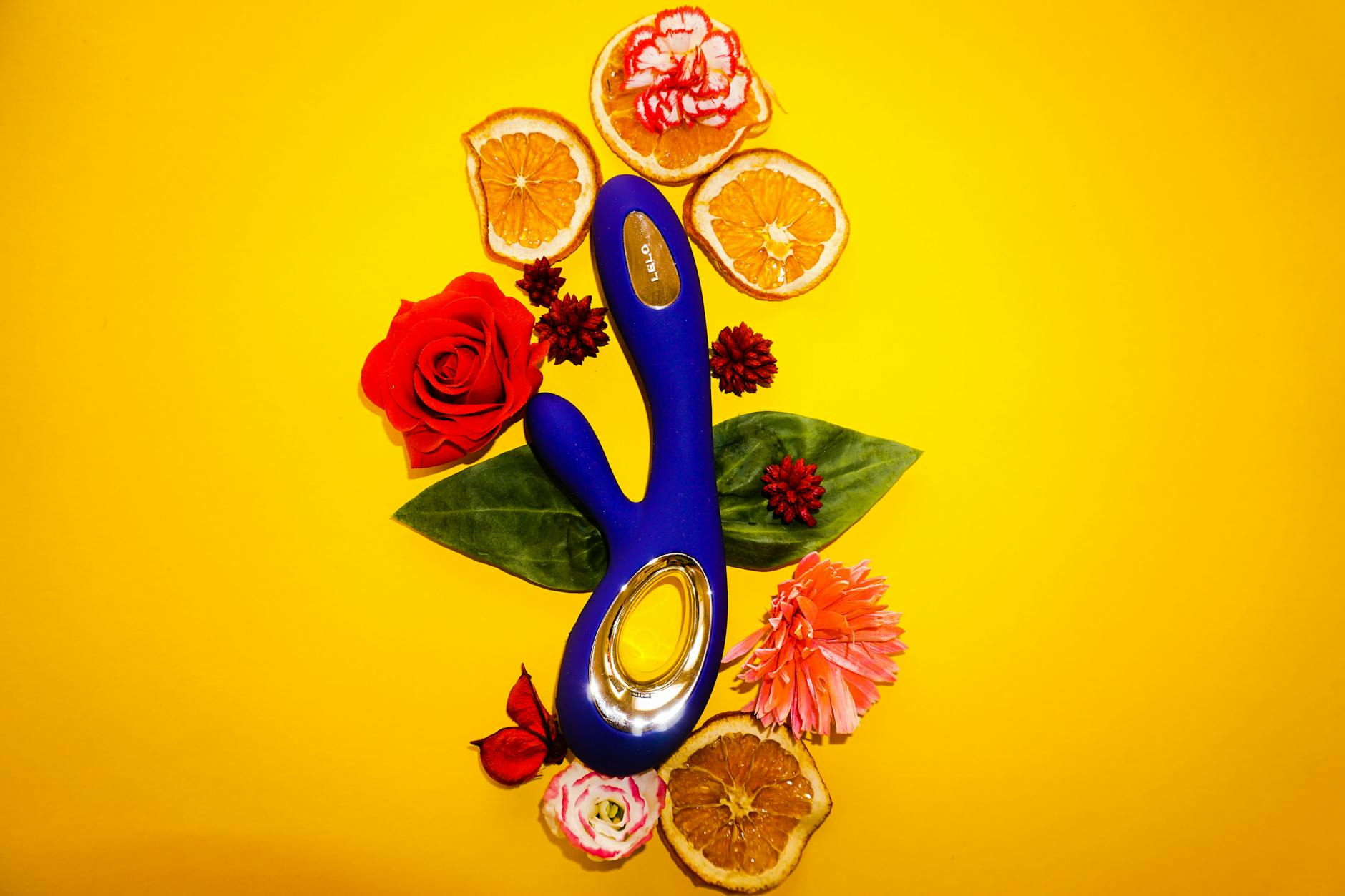 Blue vibrator surrounded by flowers and citrus on bright yellow background